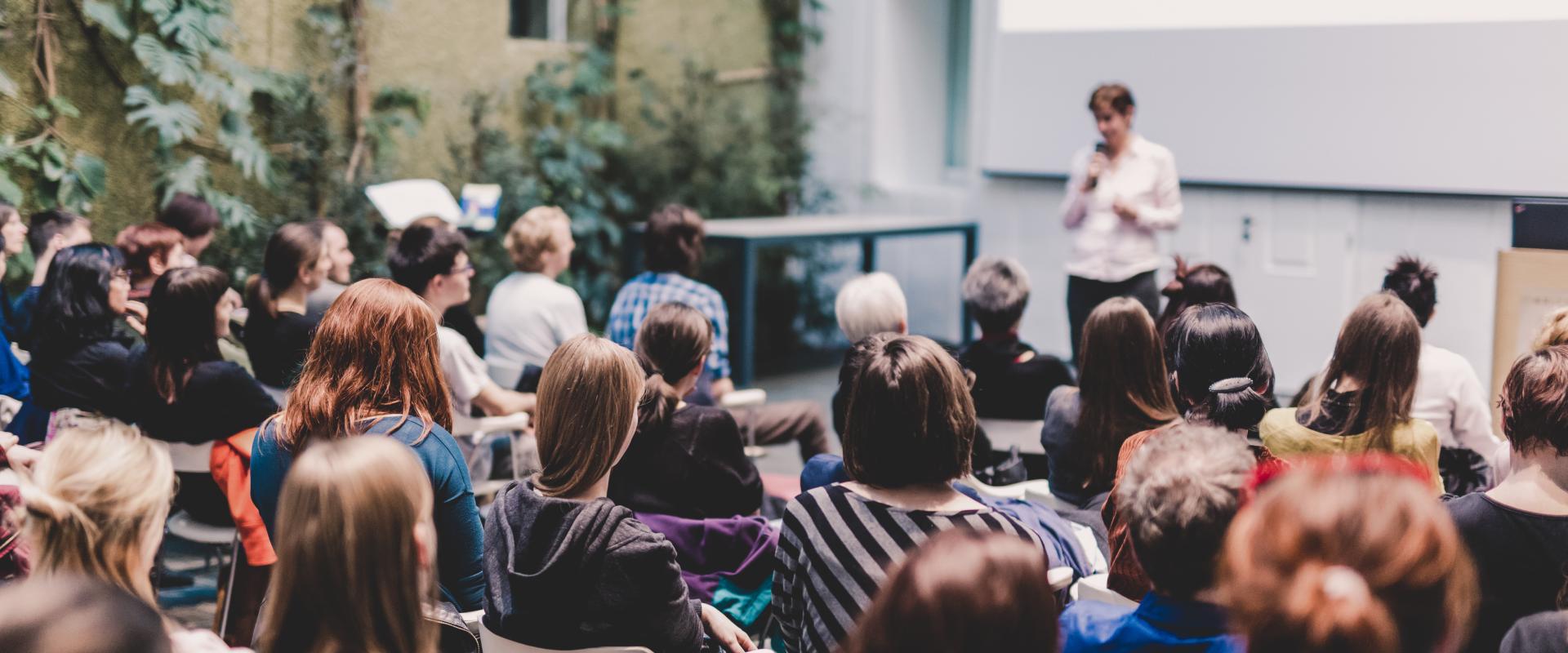 Female speaker giving presentation in lecture hall at university workshop. Audience in the conference hall. Rear view of unrecognized participant in audience. Scientific conference event